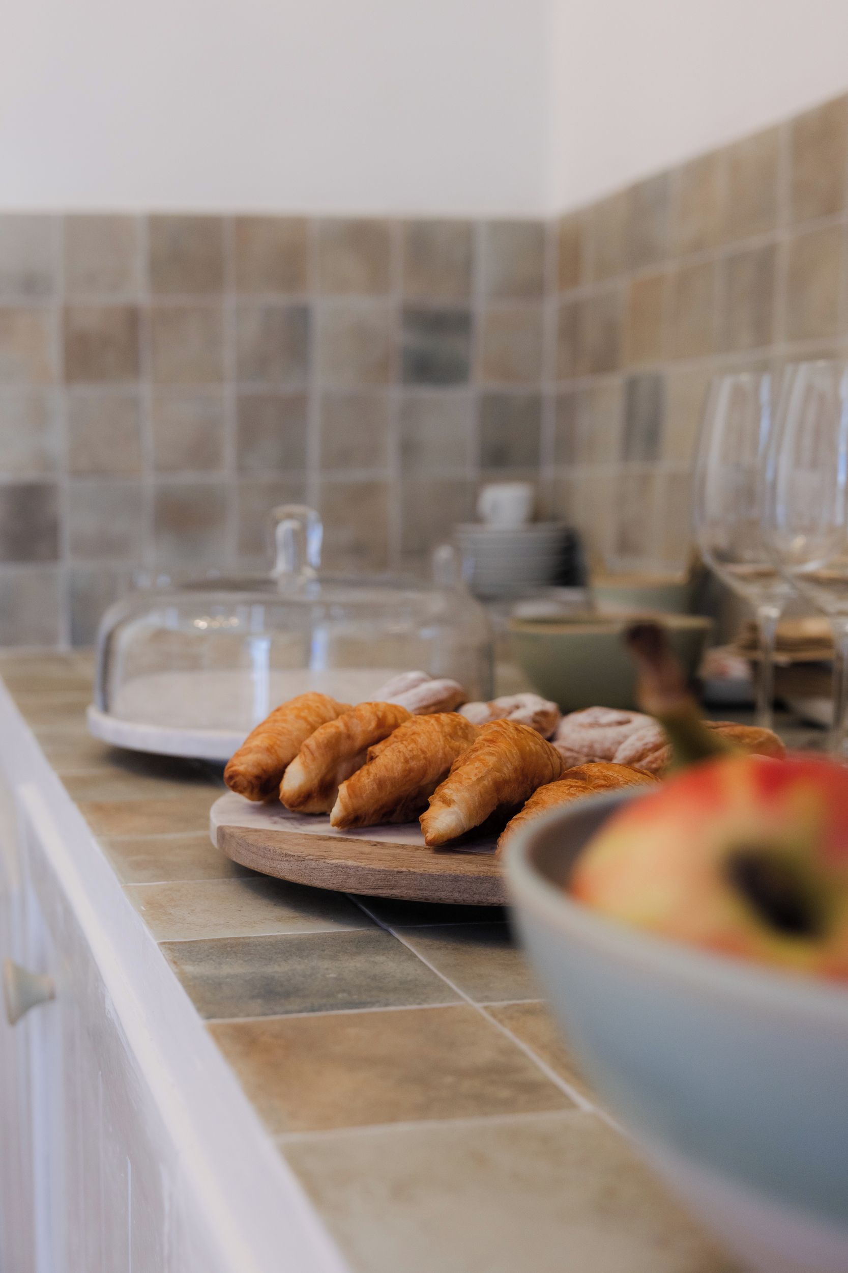 Een ontbijttafel met croissants op een houten plank, een glazen pot en een kom fruit.