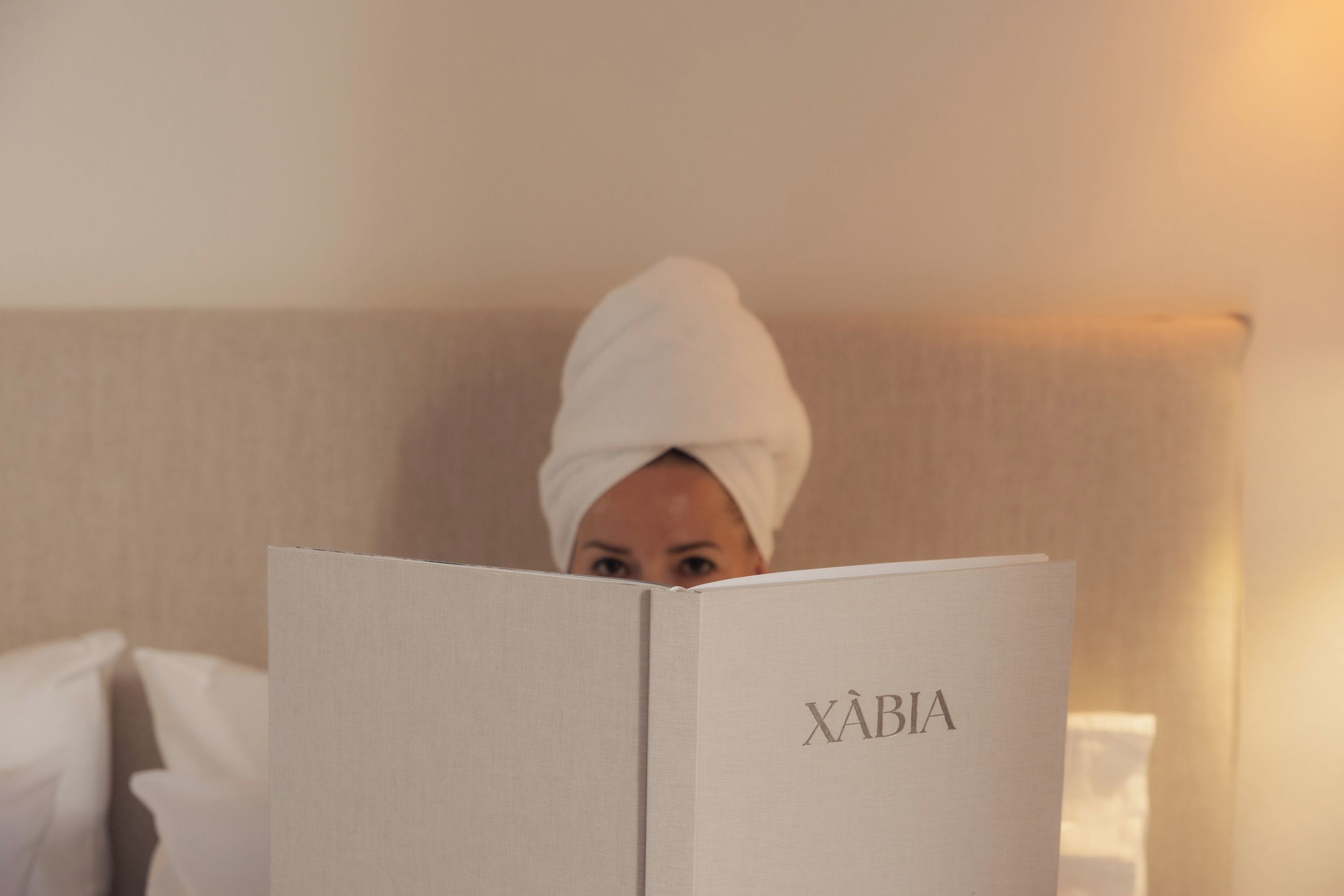 A person with a towel on their head reading a book titled 'XABIA' in a room with a bed and pillows.