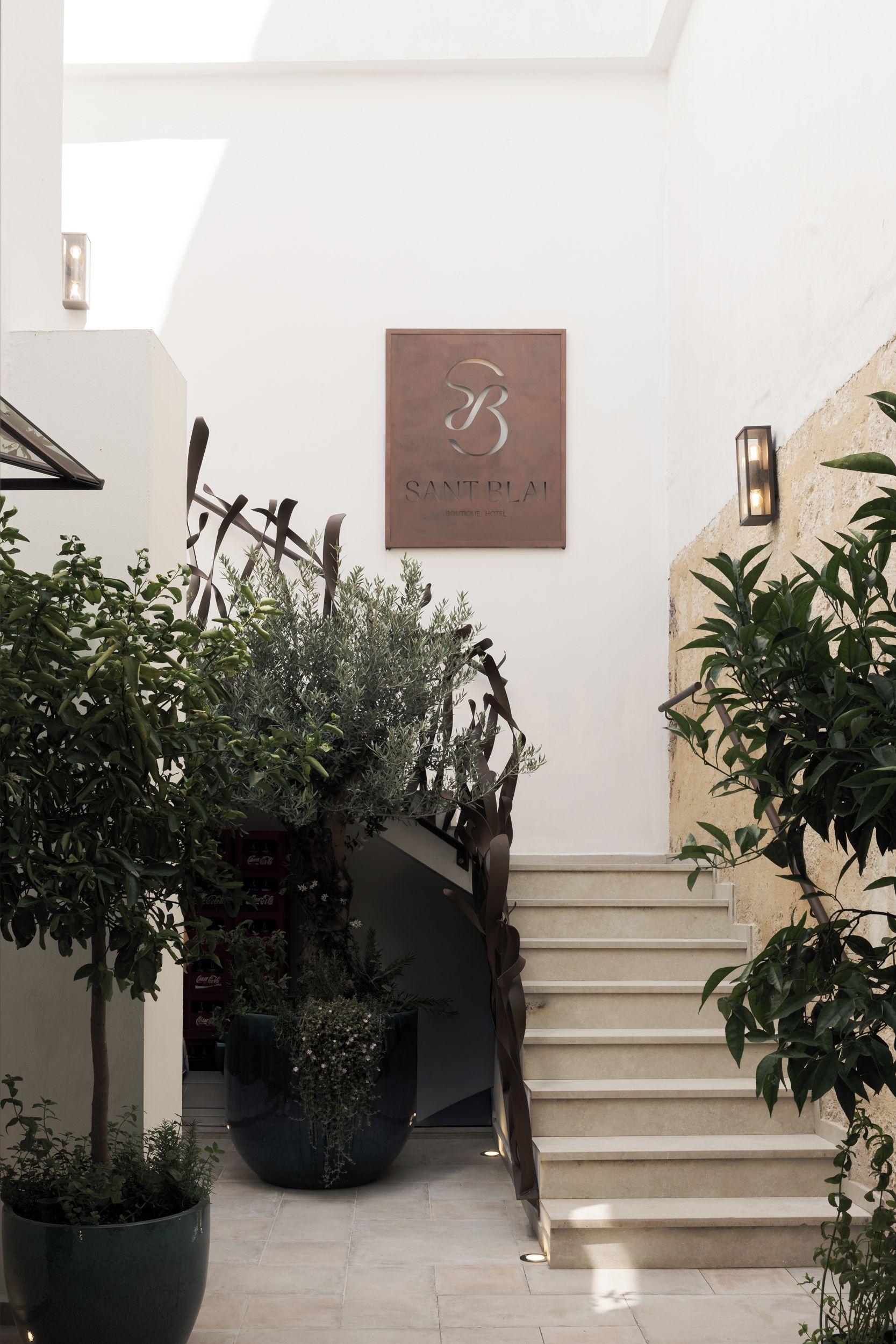 A staircase with a sign above it, surrounded by potted plants and a wall-mounted light fixture.
