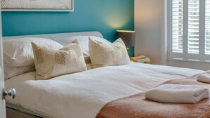 Ein ordentlich bezogenes Bett mit weißen Leinentüchern und beigen Kissen, eine türkisblaue Wand und ein Fenster mit Jalousien.