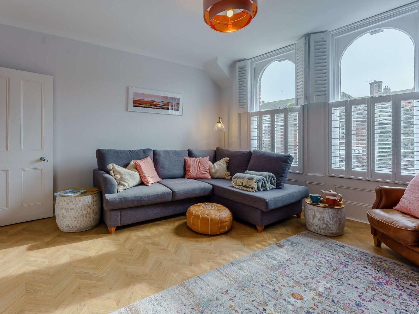 A cozy living room with a grey sectional sofa, a patterned rug, and a large window with shutters.