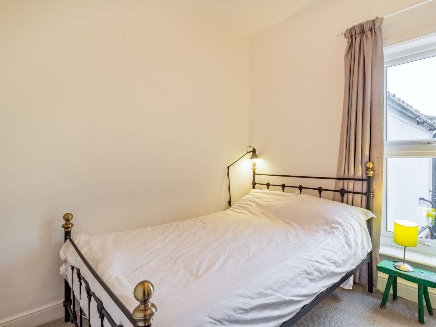Un lit simple avec une tête de lit, une lampe de chevet, une fenêtre avec des rideaux et un petit tabouret muni d’une lampe.