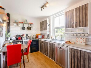 A modern kitchen with wooden cabinets, a dining area with a red chair, and a window providing natural light. - 2 Bed in Skipton (oc-87370) (Kettlewell)