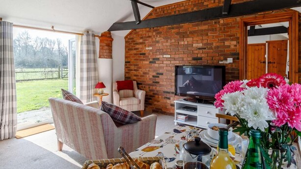 Ein gemütliches Wohnzimmer mit Backsteinwand, einem Fernseher, einem Esstisch mit Frühstücksangebot und Blick ins Freie.