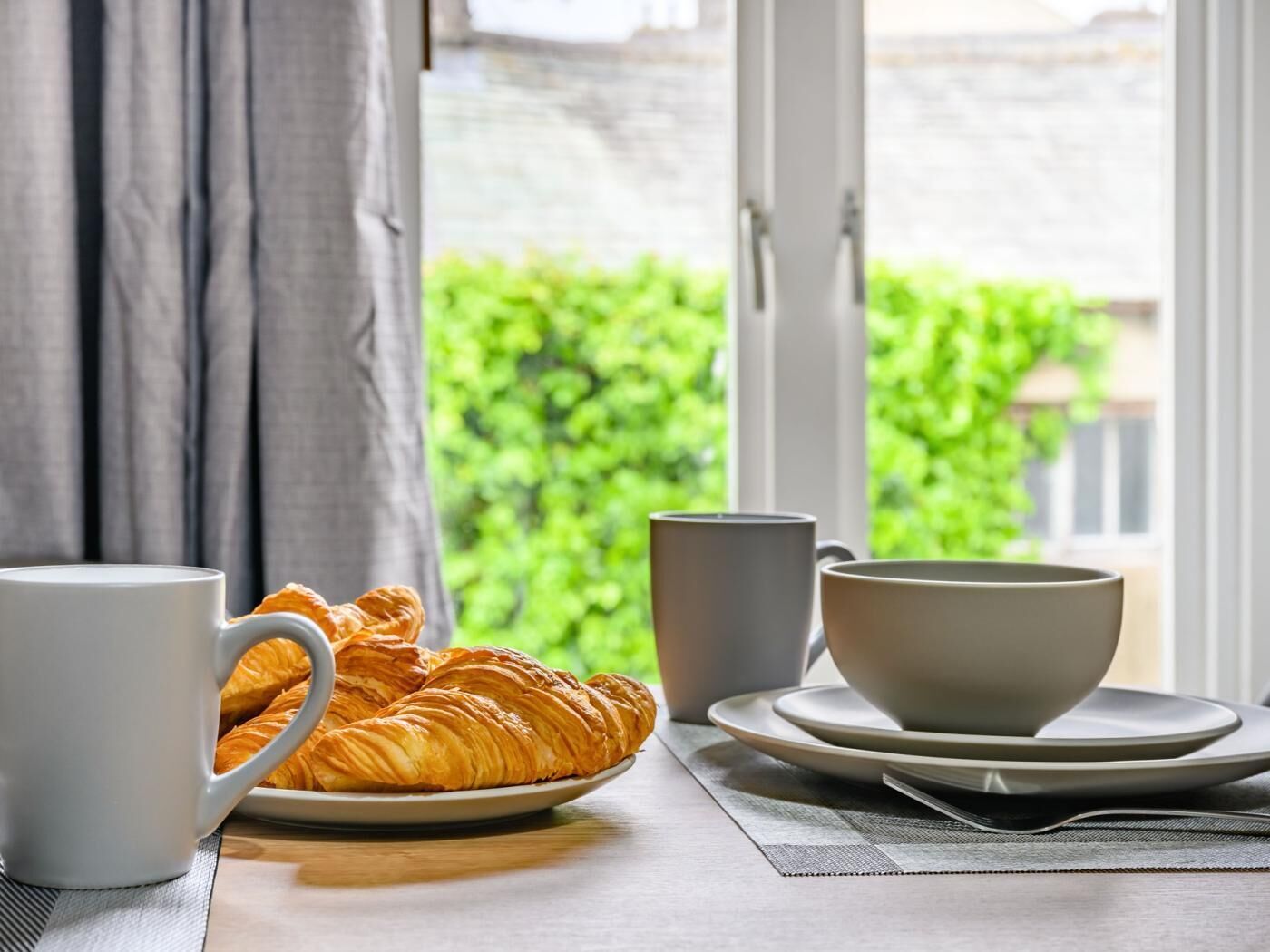 Disposição de café da manhã com croissants, uma caneca branca e uma tigela sobre uma mesa próxima a uma janela.