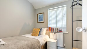Ein Schlafzimmer mit Bett, Nachttisch mit Lampe, Heizkörper, Fenster mit Jalousien und einer Wandregal mit Büchern.