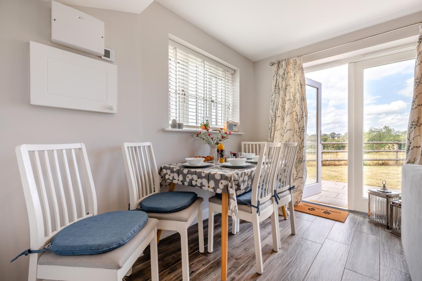 A dining area with a table set for four, white chairs with blue cushions, a window with blinds, and a view of the outdoors.