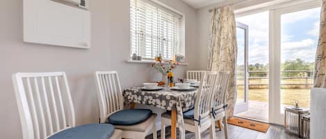 Comedor con una mesa dispuesta para cuatro personas, sillas blancas con cojines azules, una ventana con persianas y vistas al exterior.