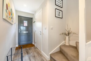 Un couloir avec un plancher en bois, une porte grise, un mur blanc orné de tableaux encadrés et un vase blanc contenant des branches.