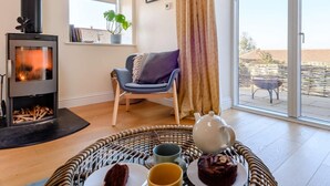 A cozy living room with a wood-burning stove, a comfortable chair, a dining table with a teapot and cake, and a view through a large sliding glass door. - 2 Bed in Norwich (oc-87511) (Colby)