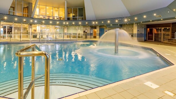 An indoor swimming pool with a central fountain, surrounded by a modern architectural design with geometric shapes and recessed lighting. - 3 Bed in Saltash (oc-87716) (Saint Mellion)