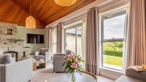 A modern living room with a stone fireplace, grey sofas, a coffee table, and large windows with curtains. - 3 Bed in Saltash (oc-87716) (Saint Mellion)