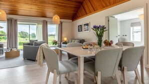A spacious living room with a wooden ceiling, a dining area with a table and chairs, and large windows offering a view of the outdoors. - 3 Bed in Saltash (oc-87716) (Saint Mellion)