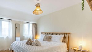 Ein Schlafzimmer mit einem hölzernen Bett, einem Nachttisch mit Lampe, einem Fenster mit Vorhängen und einer hängenden Pflanze.
