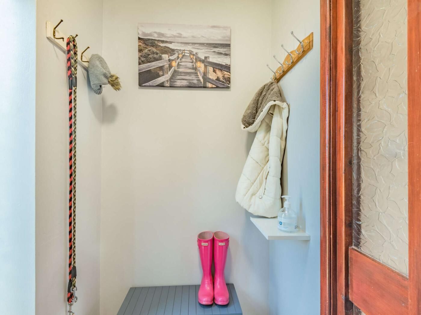 Une pièce avec un banc en bois, un porte-manteau, une serviette suspendue, une photo d’un sentier côtier et des bottes en caoutchouc roses.