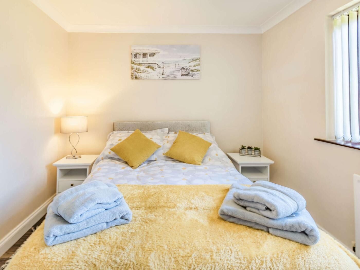 A bedroom with a bed, two nightstands, a lamp, a painting, and towels.
