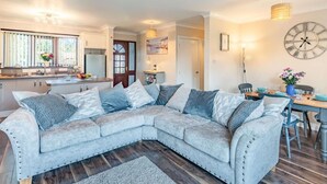A modern living room with a sectional sofa, a dining table with chairs, a kitchen area, and a large wall clock. - 2 Bed in Mablethorpe (oc-87801) (Mablethorpe)