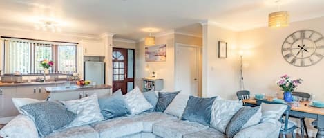 En moderne stue med en hjørnesofa, et spisebord med stole, et køkkenområde og et stort vægur.
