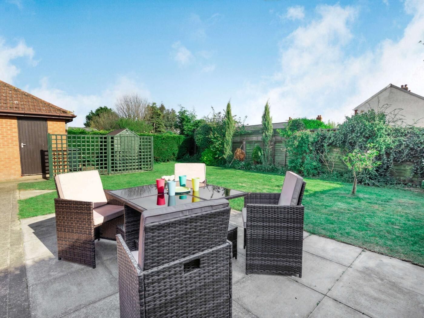 An outdoor patio with wicker furniture, a glass-top table, and a green lawn.