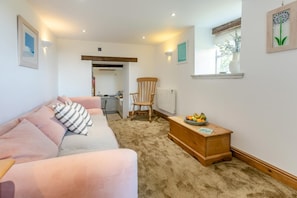 A cozy living room with a sofa, a wooden side table with fruit, a chair, and a view of a kitchen and a window with a plant. - 1 Bed in Penzance (oc-88008) (Longrock)