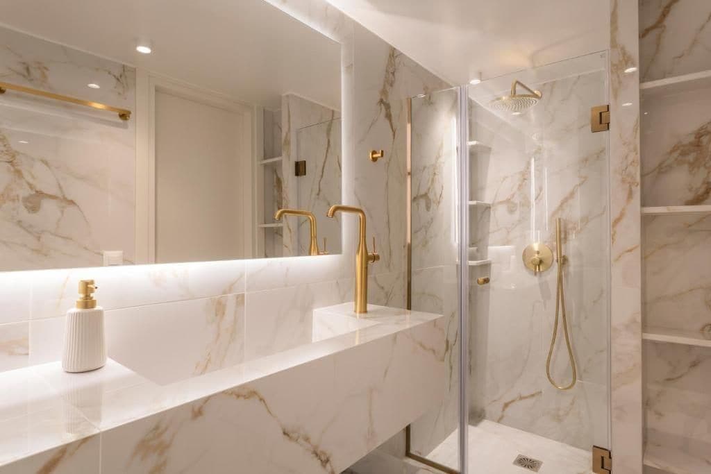 A modern bathroom with a marble countertop, gold fixtures, and a glass-enclosed shower.