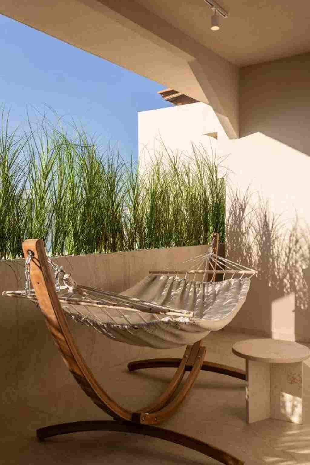 A hammock chair with a metal frame and a fabric seat, positioned on a balcony with a view of tall grass and a building in the background.