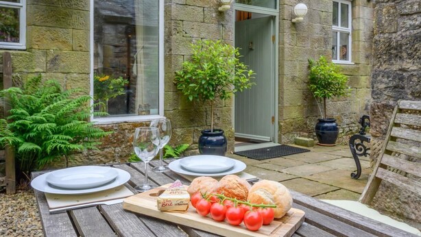 A stone building with a wooden table set for a meal, including bread, tomatoes, and a bottle of wine. - 2 Bed in Hexham (oc-88032) (Once Brewed)