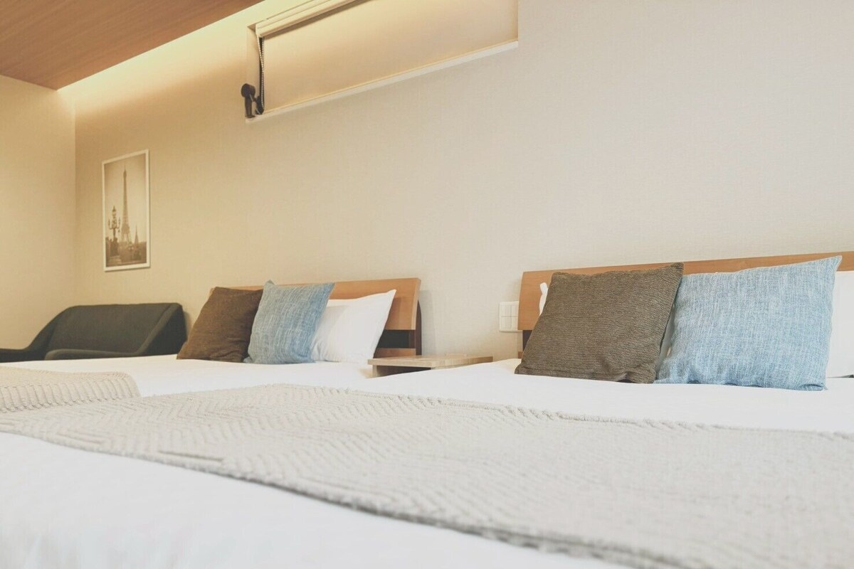 A hotel room with two beds, a chair, a bedside table, and a wall-mounted light fixture.