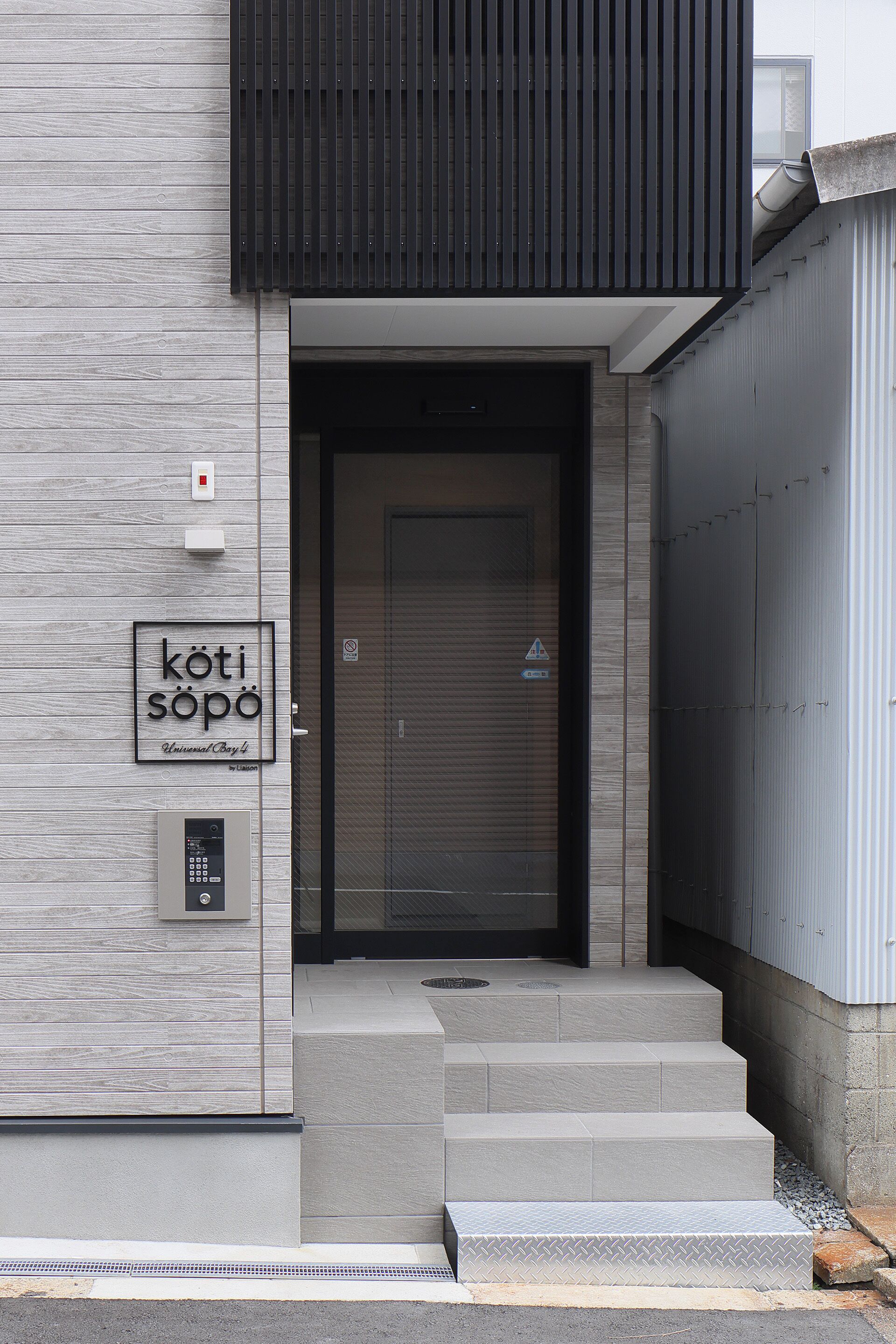 A modern building entrance with a metal door, steps, and a sign that reads 'köti sopo'.