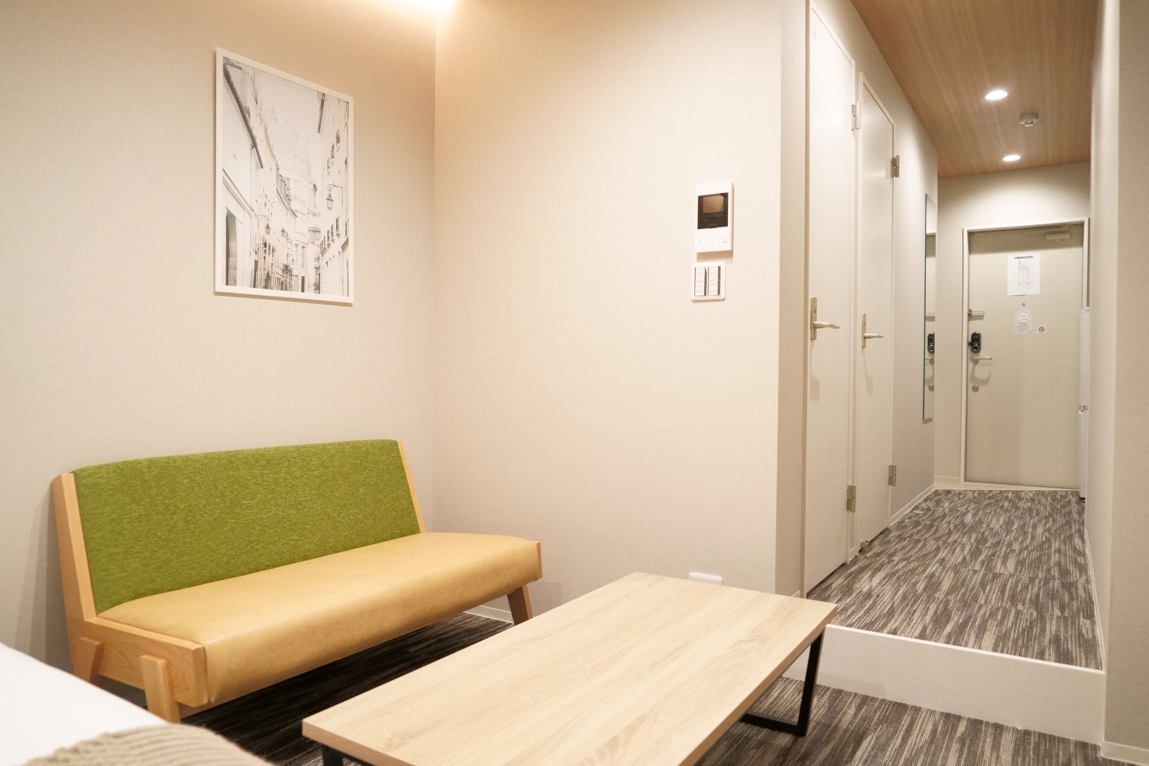 Une chambre d’hôtel avec un banc rembourré de tissu vert, une table en bois, un thermostat fixé au mur et une porte dotée d’un panneau en verre.