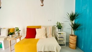 Een bed met een geel hoofdeinde, een rood kussen en een witte deken. Op een nachtkastje naast het bed staan twee potten met planten.