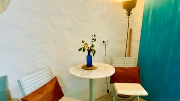 Een kleine kamer met een witte muur, een ronde tafel met een blauwe vaas, twee witte stoelen met bruine kussens en een hoge vloerlamp.