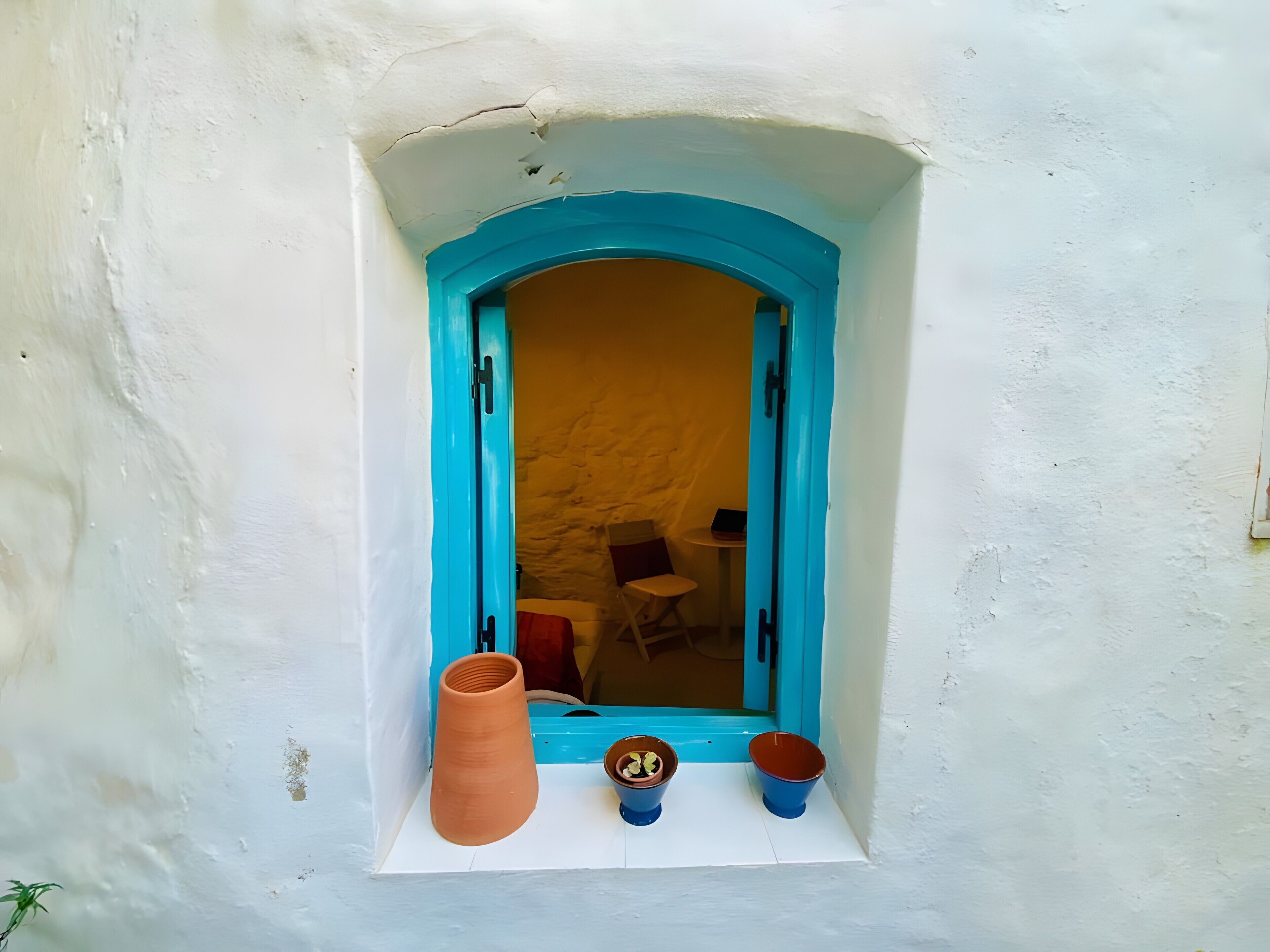 Uma janela com moldura azul, uma parede branca e uma pequena estante com um vaso de terracota e duas tigelas azuis.
