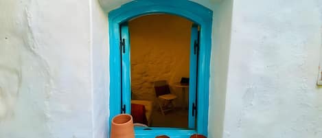 Uma janela com moldura azul, uma parede branca e uma pequena estante com um vaso de terracota e duas tigelas azuis.