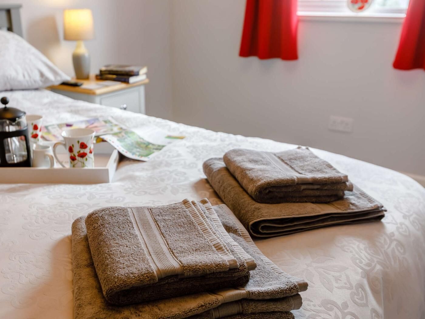 Una cama bien hecha con toallas dobladas, una mesita de noche con una lámpara y una ventana con cortinas rojas.