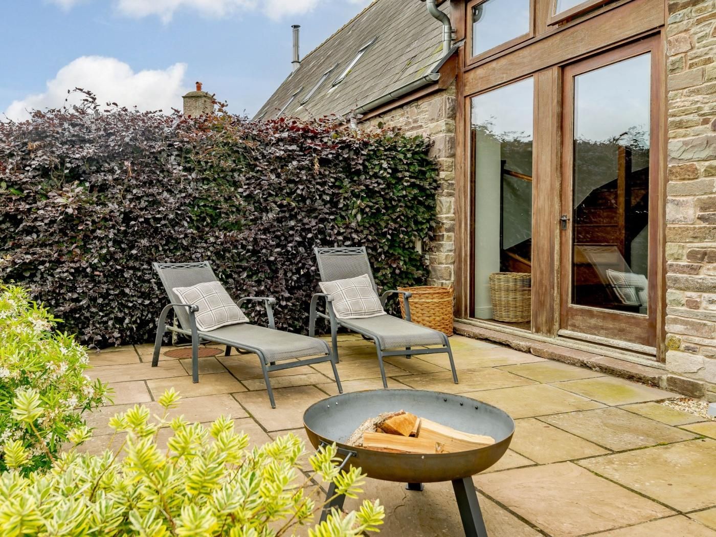 A patio with two lounge chairs, a fire pit, and a stone wall with climbing plants.