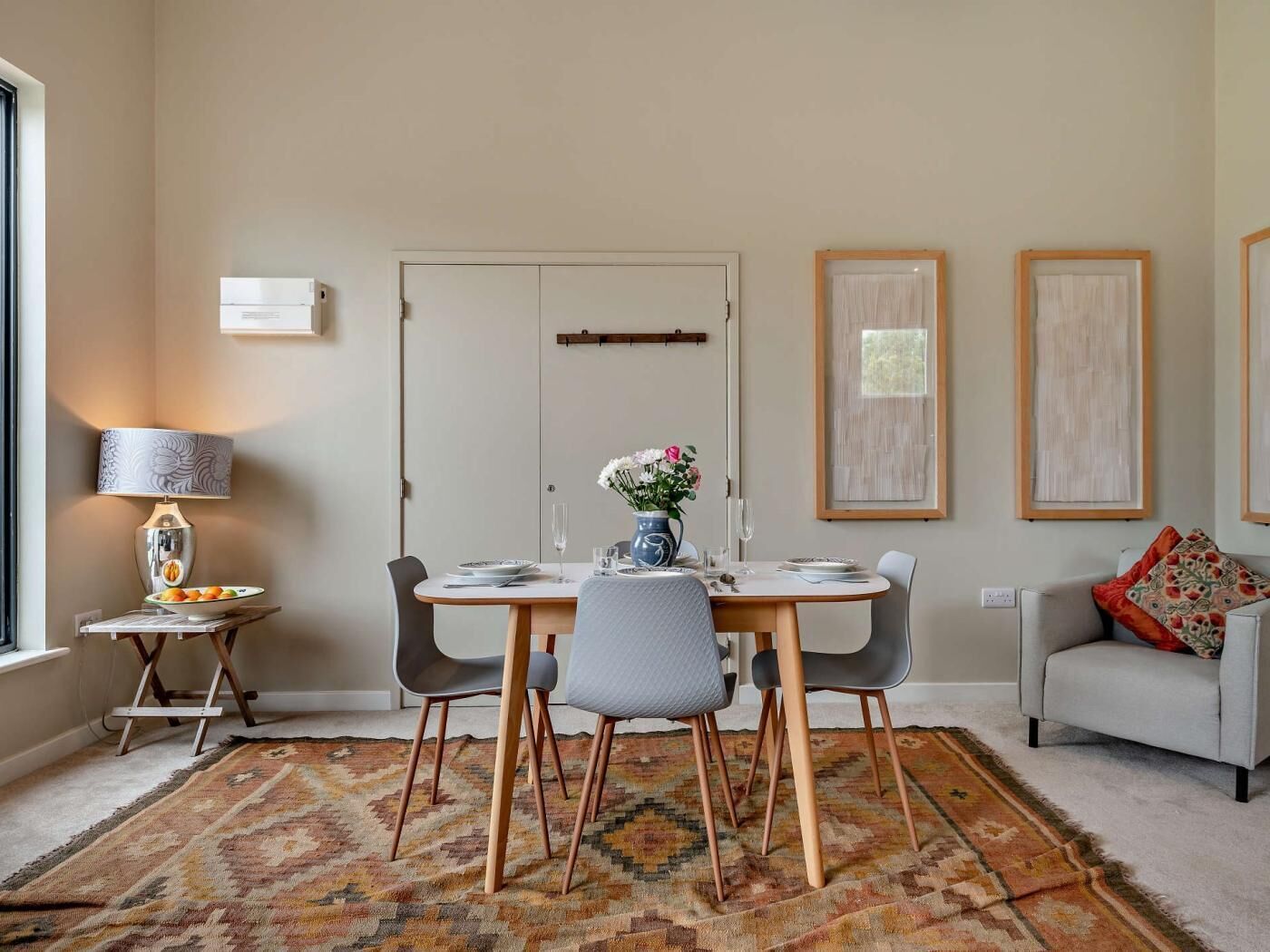 A dining area with a table set for four, a lamp, a vase with flowers, and framed artwork on the wall.