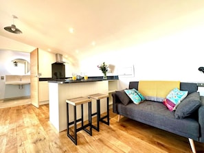 A modern kitchen with a central island, a grey sofa with colorful cushions, and a wooden floor.