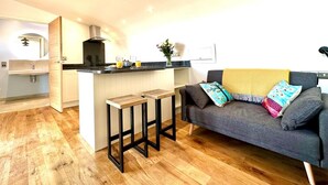 Eine moderne Küche mit einer zentralen Kochinsel, ein graues Sofa mit bunten Kissen und ein Holzboden.