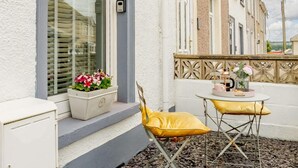 Ein Balkon mit einem weißen Schrank, einem gelben Stuhl, einem kleinen Tisch mit Tasse und Blumen sowie einem Fenster mit Jalousien.