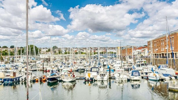 Ein Yachthafen mit zahlreichen vertäuten Booten, blauem Himmel und Gebäuden im Hintergrund.