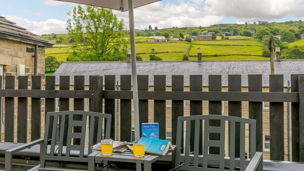 En terrasse med udsigt over grønne marker og huse, et bord med drikkevarer og en bog, samt et træhegn.