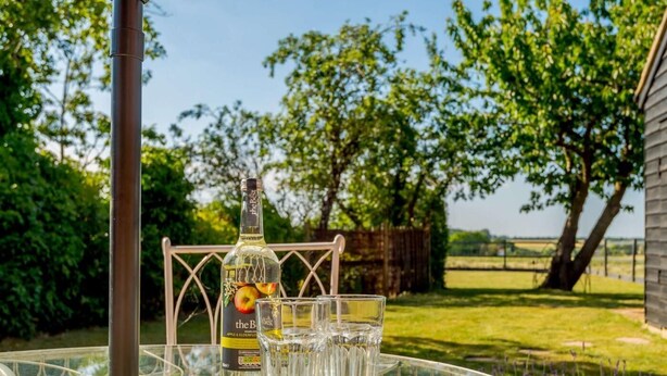 Ett glasbord med en flaska och glas, placerat utomhus tillsammans med en metallstol, omgivet av grönska och ett staket.