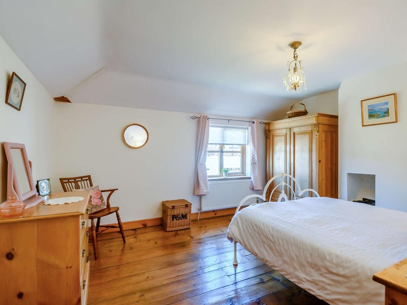 Een slaapkamer met een houten vloer, een bed, een dressoir, een stoel, een raam met gordijnen, een spiegel en een kroonluchter.