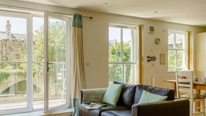 A cozy living room with a leather sofa, a dining table, and large windows offering a view of greenery and a building. - 2 Bed in Tenby (oc-88738) (Tenby)