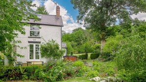 A white house with a chimney, a bench, and a garden with various plants and a small pond. - 4 Bed in Corwen (oc-88868) (Corwen)