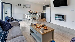 A modern living room with a sofa, a coffee table with croissants, a dining area with chairs, a kitchen with a refrigerator, and a TV mounted on the wall. - 2 Bed in Camborne (oc-88826) (Camborne)