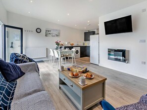 Ein modernes Wohnzimmer mit einer Couch, einem Couchtisch mit Croissants, einem Essbereich mit Stühlen, einer Küche mit Kühlschrank und einem an der Wand montierten Fernseher.