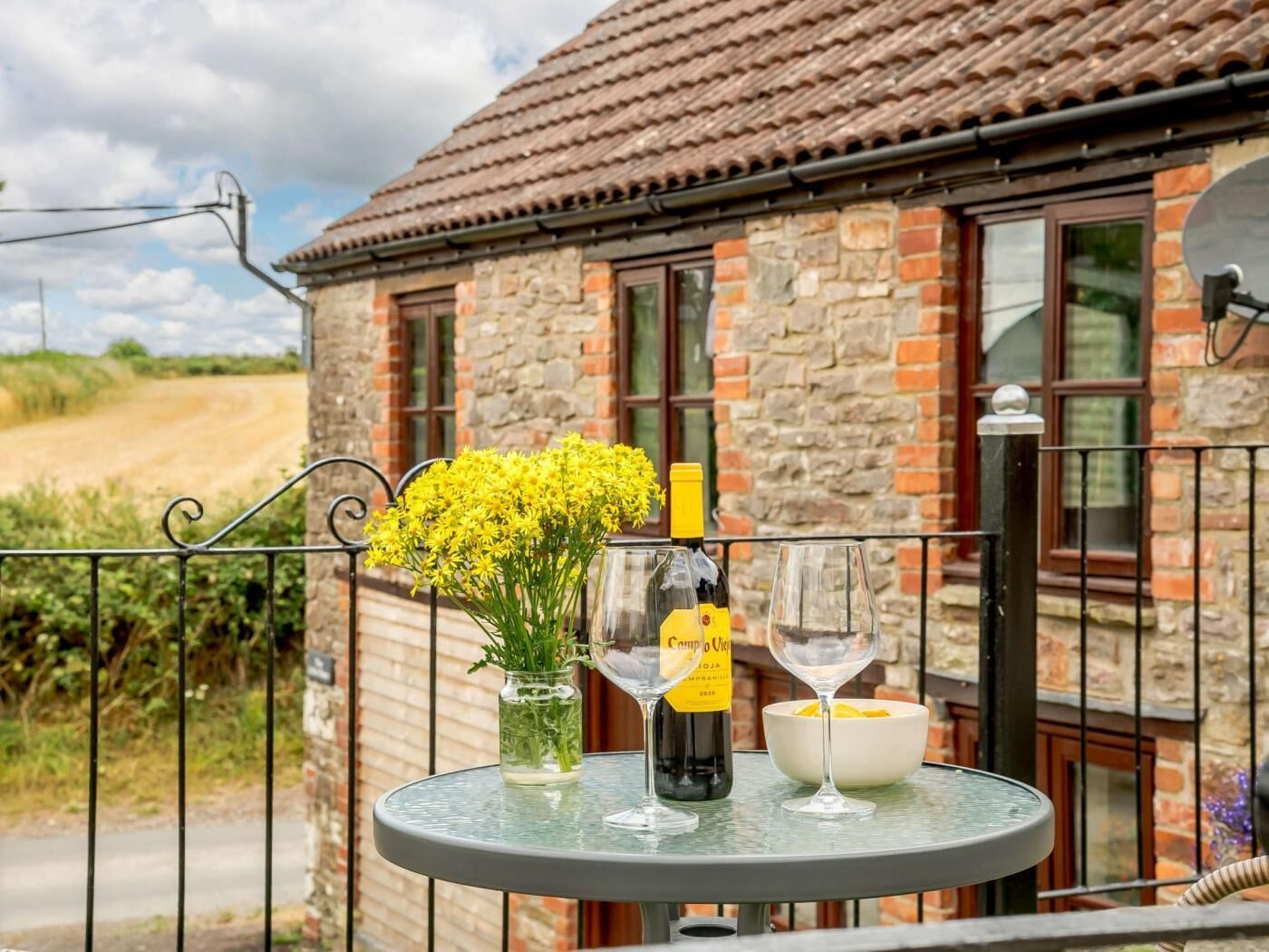 A balcony with a table set for two, featuring a bottle of wine, a glass, and a vase with yellow flowers.
