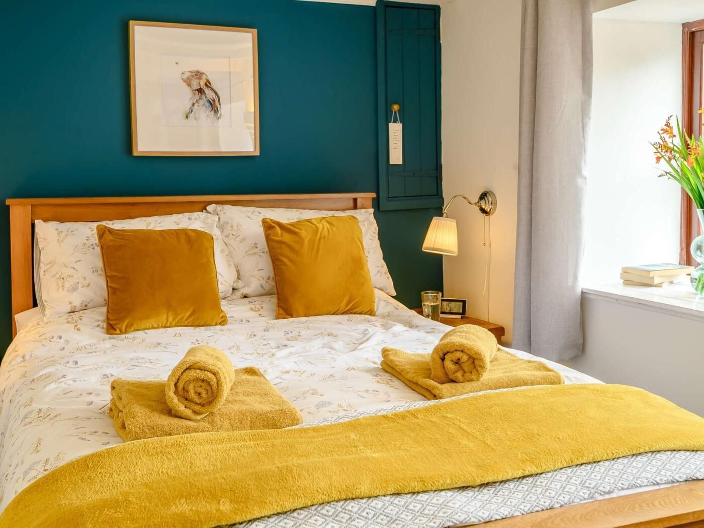 Een net opgemaakt bed met een beddegoed met bloemenprint, twee opgerolde handdoeken en een bedlamp.